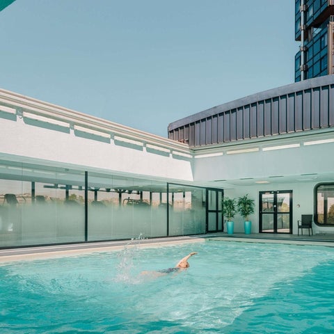 A swimmer is in a clear blue indoor pool with a modern building and glass walls visible in the background.