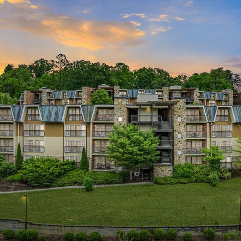 A multi-story building with a stone facade and balconies overlooks a manicured lawn and lush greenery at dusk.