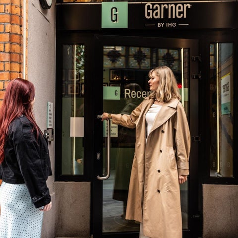 Two people stand outside the entrance of the Garner by IHO hotel, with a brick facade and glass doors.