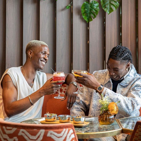 Two people toast with cocktails while seated at a table with patterned upholstery and decorative accents in a stylish bar.