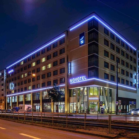 A modern Novotel hotel building is illuminated with blue and orange lights at night, featuring a prominent logo and street-level entrance.