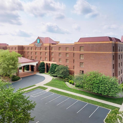 A large, brick hotel with a covered entrance and ample parking is surrounded by lush greenery and trees.