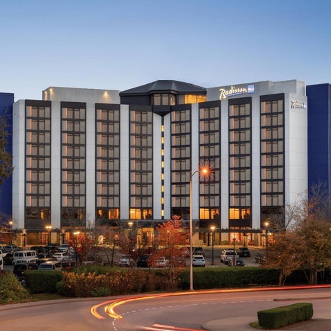 A Radisson hotel building facade is shown at dusk, featuring a modern design with blue and white accents and illuminated windows.