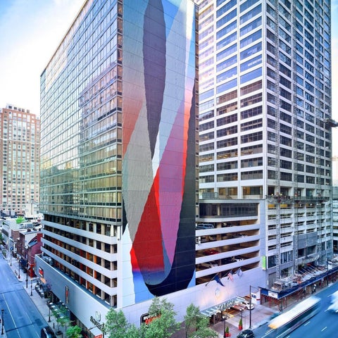 A modern building with a large, colorful abstract mural on its facade, situated on a city street with traffic and surrounding buildings.