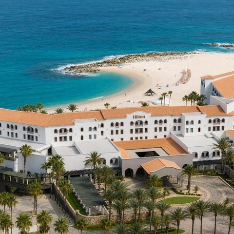 An aerial view showcases a large, white hotel with red-tiled roofs and palm trees bordering a sandy beach and turquoise ocean.