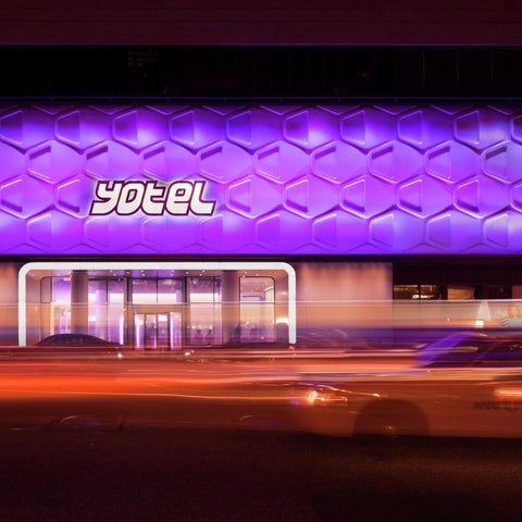 A brightly lit hotel facade features a hexagonal pattern and the 'yotel' logo, with blurred car lights in the foreground.