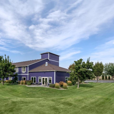 A Best Western Plus hotel building with a blue and purple facade stands on a well-manicured lawn under a partly cloudy sky.