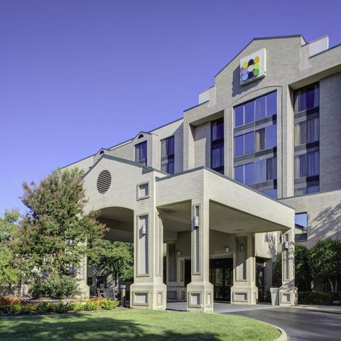 A large, multi-story hotel building with a covered entrance and landscaping is shown on a sunny day.
