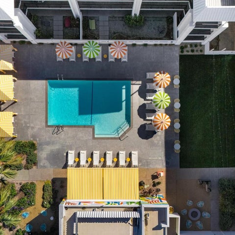 An aerial view showcases a rectangular swimming pool surrounded by colorful umbrellas, lounge chairs, and lush landscaping.